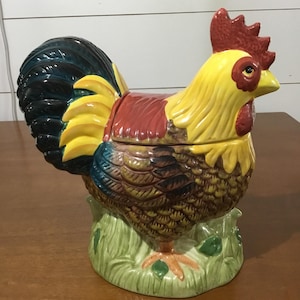 May include: A ceramic rooster-shaped cookie jar. The jar is painted in bright colours, including yellow, red, brown, and green. The rooster stands on a green base with grass and leaves. The tail feathers are black and blue.