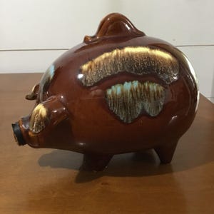 Pode incluir: Um cofrinho de cerâmica marrom com acabamento brilhante. O banco tem um corpo arredondado, pernas pequenas e um focinho. Possui detalhes decorativos em bege, amarelo e azul claro. O cofrinho é um item decorativo clássico.