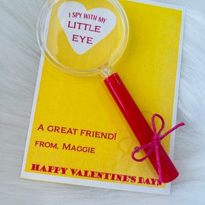 May include: A yellow card with the text "I spy with my little eye" inside a red heart shape. A magnifying glass with a red handle is attached to the card with a pink ribbon. The card also says "A great friend! From, Maggie" and "Happy Valentine's Day!"
