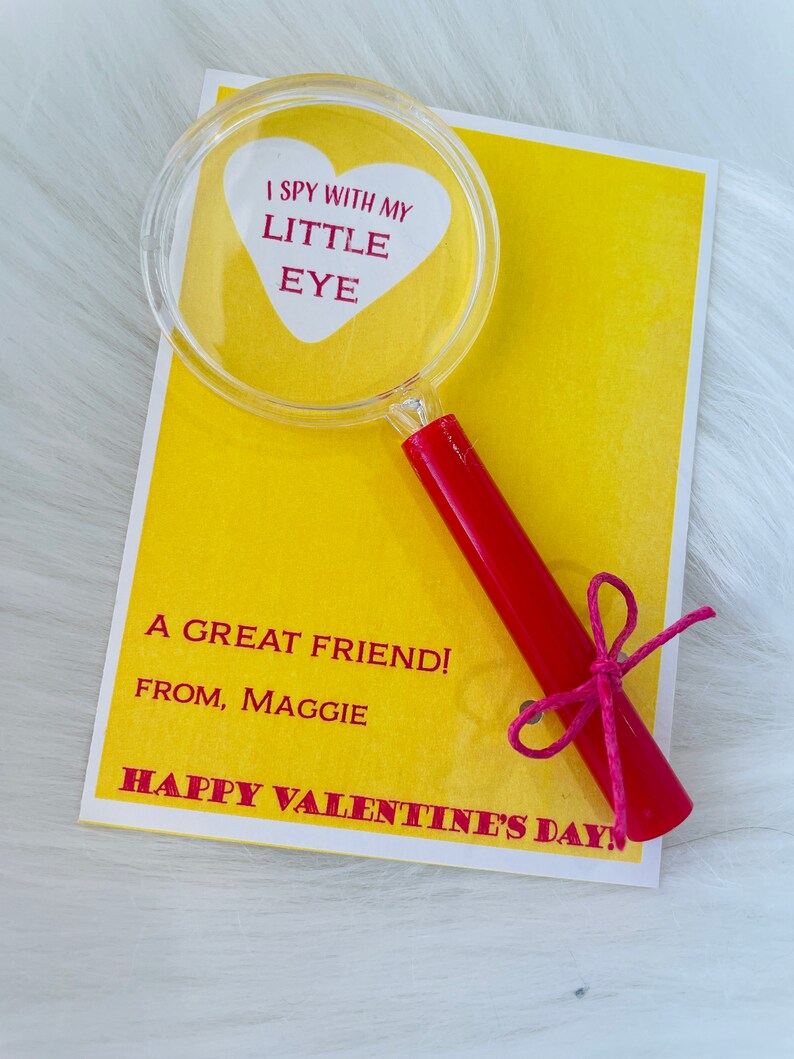 May include: A yellow card with the text "I spy with my little eye" inside a red heart shape. A magnifying glass with a red handle is attached to the card with a pink ribbon. The card also says "A great friend! From, Maggie" and "Happy Valentine's Day!"