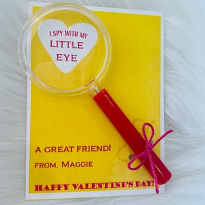 May include: A yellow card with the text "I spy with my little eye" inside a red heart shape on a magnifying glass with a red handle tied with a pink ribbon. The card also says "A great friend! From, Maggie Happy Valentine's Day!"