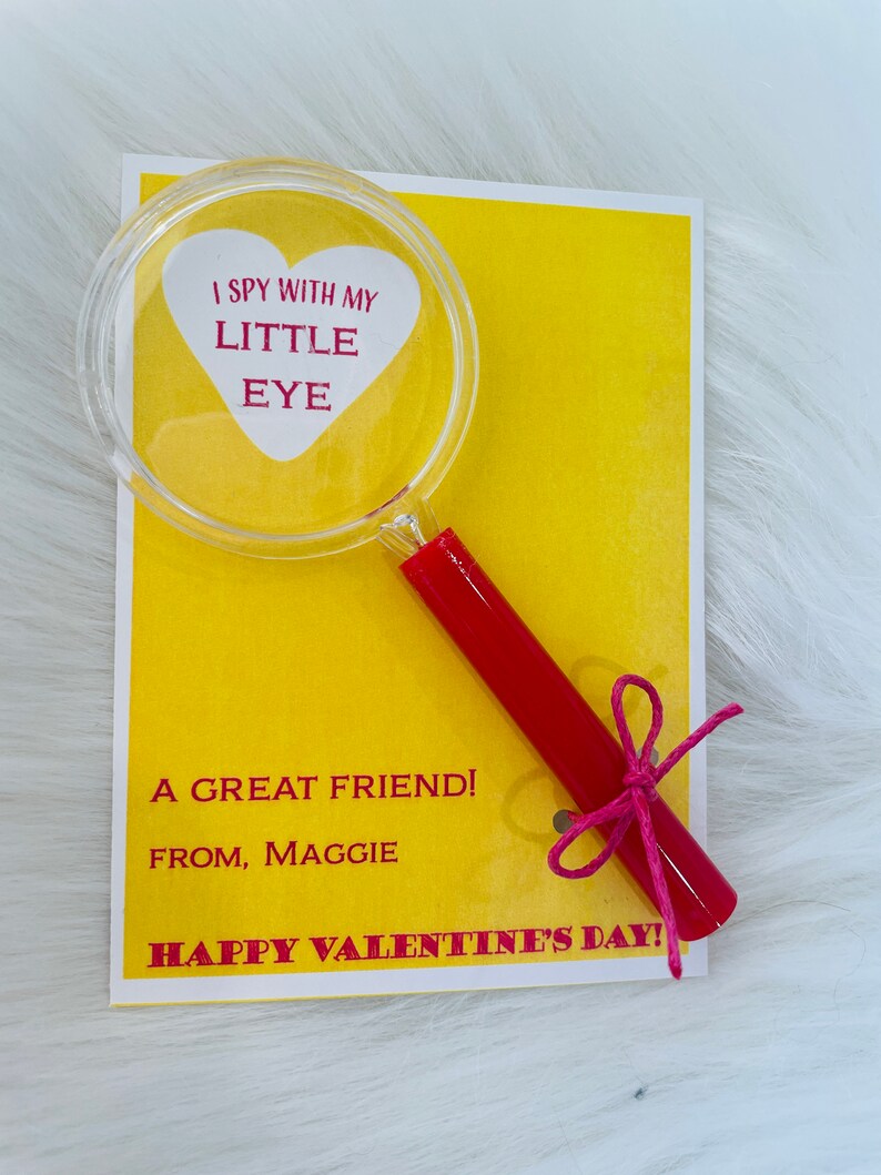 May include: A yellow card with the text "I spy with my little eye" inside a red heart shape on a magnifying glass with a red handle tied with a pink ribbon. The card also says "A great friend! From, Maggie Happy Valentine's Day!"