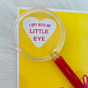 May include: A yellow card with a red heart-shaped magnifying glass on top. The magnifying glass has the text "I spy with my little eye" inside the heart. The card also has the text "A great friend! From, Maggie" on it.