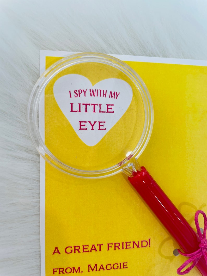 May include: A yellow card with a red heart-shaped magnifying glass on top. The magnifying glass has the text "I spy with my little eye" inside the heart. The card also has the text "A great friend! From, Maggie" on it.