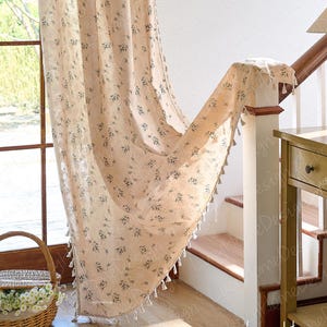 Op de afbeelding: Beige gordijn met bloemenpatroon en witte kwastjes. Het gordijn hangt voor een raam en een trap. Een rieten mand met bloemen en een rieten lamp zijn ook zichtbaar.