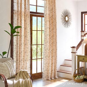 Op de afbeelding: Beige gordijnen met bloemenpatroon hangen voor een houten deur en raam. De gordijnen hebben een delicaat bloemmotief en worden gecombineerd met een bijpassende deken en decoratief kussen op een stoel. Een zonne-spiegel hangt aan de muur.