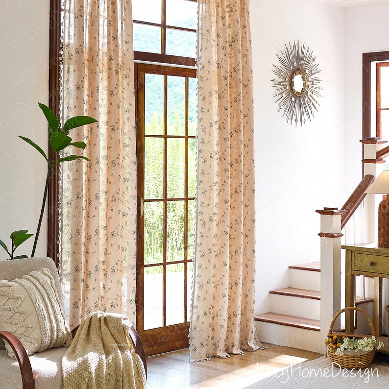 Op de afbeelding: Beige gordijnen met bloemenpatroon hangen voor een houten deur en raam. De gordijnen hebben een delicaat bloemmotief en worden gecombineerd met een bijpassende deken en decoratief kussen op een stoel. Een zonne-spiegel hangt aan de muur.