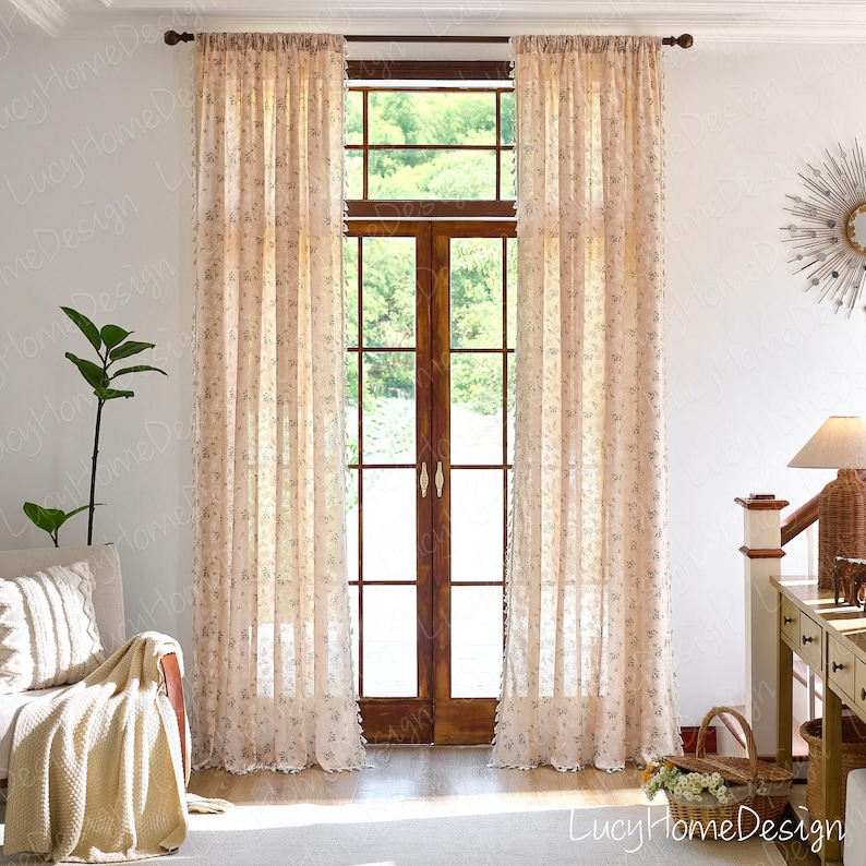 Op de afbeelding: Transparante beige bloemengordijnen die een houten raamkozijn omlijsten. De gordijnen hebben een delicaat bloemenpatroon en hangen aan een donkerbruine stang. Een bank met een cr&egrave;mekleurige plaid en een plant zijn zichtbaar.