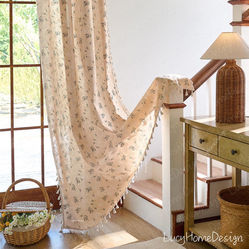 Op de afbeelding: Beige gordijn met bloemenpatroon en witte kwastjes. Het gordijn hangt voor een raam en een trap. Een rieten mand met bloemen en een rieten lamp zijn ook zichtbaar.