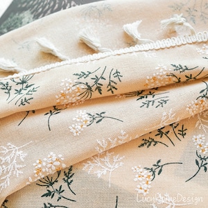 Op de afbeelding: Close-up van beige stof met een bloemmotief van witte en gele bloemen en groene bladeren. De stof heeft een witte rand met kwastjes. De textuur en het bloemmotief worden uitgelicht.
