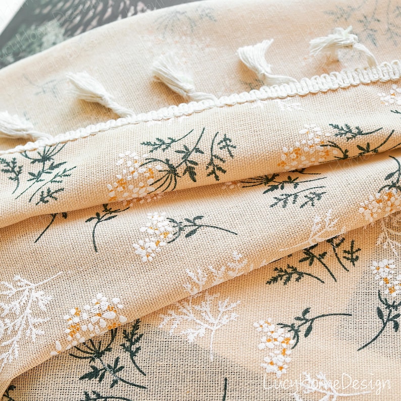 Op de afbeelding: Close-up van beige stof met een bloemmotief van witte en gele bloemen en groene bladeren. De stof heeft een witte rand met kwastjes. De textuur en het bloemmotief worden uitgelicht.