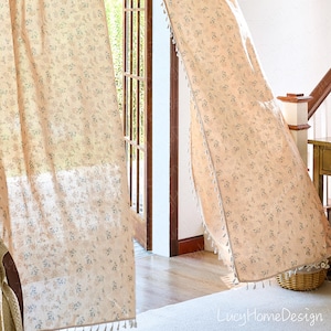 Op de afbeelding: Transparante beige gordijnen met een delicaat bloemmotief in blauw en roze. De gordijnen zijn afgezet met kwastjes langs de randen en hangen voor een deuropening. De tekst "LucyHomeDesign" is rechtsonder zichtbaar.