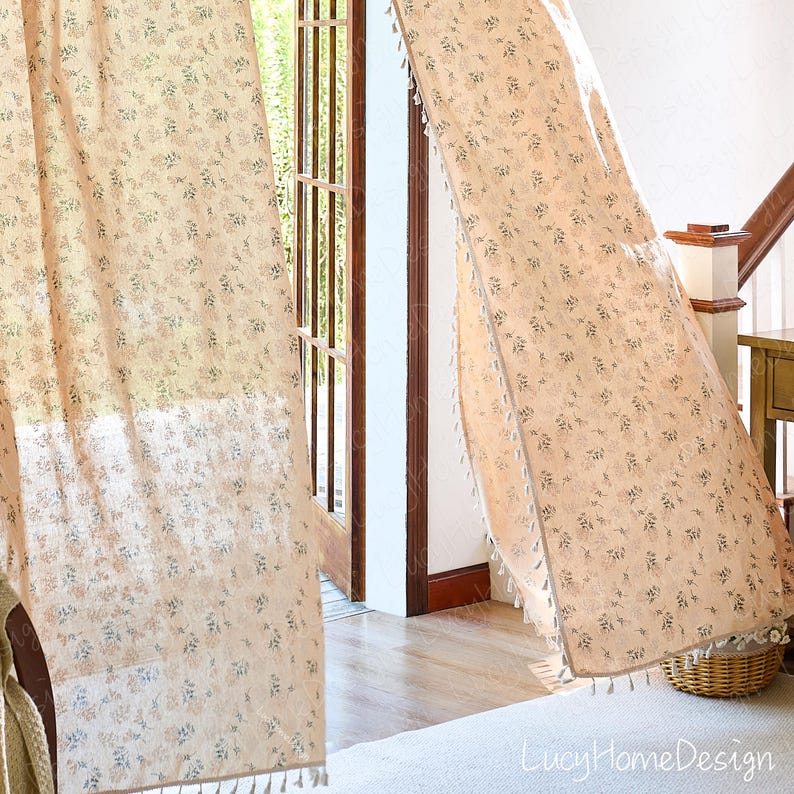 Op de afbeelding: Transparante beige gordijnen met een delicaat bloemmotief in blauw en roze. De gordijnen zijn afgezet met kwastjes langs de randen en hangen voor een deuropening. De tekst "LucyHomeDesign" is rechtsonder zichtbaar.