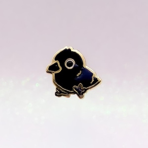 May include: A small enamel pin shaped like a stylised black bird with a gold-coloured metal outline. The bird features a single white eye with a black pupil, a small beak, and a wing detail, all rendered in a simple, flat graphic design style.