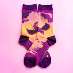 Könnte beinhalten: Ein Paar wadenlange Socken mit einem wiederkehrenden Muster aus violetten Drachen, die vor einem sonnenuntergangsartigen Hintergrund in Orange-, Gelb- und Rosatönen fliegen. Das Design enthält kleine gelbe Sternakzente sowie violette Bündchen, Fersen und Zehen.
