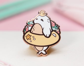May include: A rose gold enamel pin featuring a cartoon seal wearing a yellow life preserver with flowers and a starfish. The seal is wearing a yellow duck on its head.