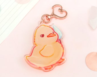 May include: A clear acrylic charm featuring a cartoon duckling with a light orange body, red accents, and a blue hat. The charm has a rose gold heart-shaped clasp. The background is light pink with rose gold confetti.