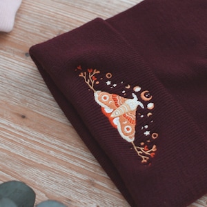 May include: A burgundy beanie with an embroidered design of a moth with wings spread, surrounded by stars and crescent moons. The moth is orange and yellow with black accents.