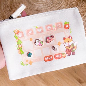 Op de afbeelding: Witte canvas pouch met een ritssluiting en een grillig ontwerp. Het ontwerp toont een raster met verschillende pictogrammen, waaronder een voskarakter, en de woorden "DROP" en "USE" in het rood. Decoratieve spelden zijn ook zichtbaar.