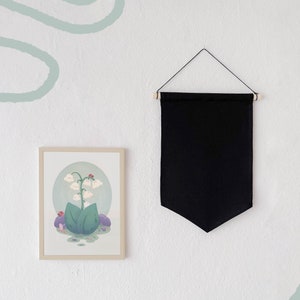 Op de afbeelding: Een ingelijste illustratie van een groene plant met witte bloemen en rode lieveheersbeestjes. De illustratie is op een witte achtergrond en hangt aan een witte muur met een zwarte banner erboven.