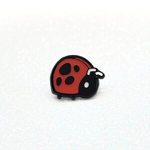 Puede incluir: Un pequeño pin de mariquita roja con manchas negras y contorno negro. El pin está sobre una superficie blanca.
