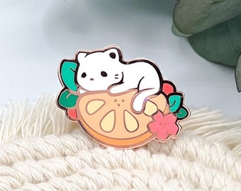 May include: White enamel pin of a cat sleeping on a slice of orange with green leaves and a pink flower.