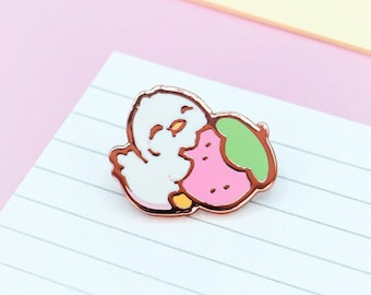 May include: A cute enamel pin featuring a white chick hugging a pink strawberry with a green stem. The pin has a rose gold outline and is set on a lined white paper. The background is a soft pink.