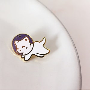 Puede incluir: Pin de esmalte dorado con un gato blanco que lleva un casco de astronauta morado. El gato est&aacute; en posici&oacute;n flotante con las patas extendidas.