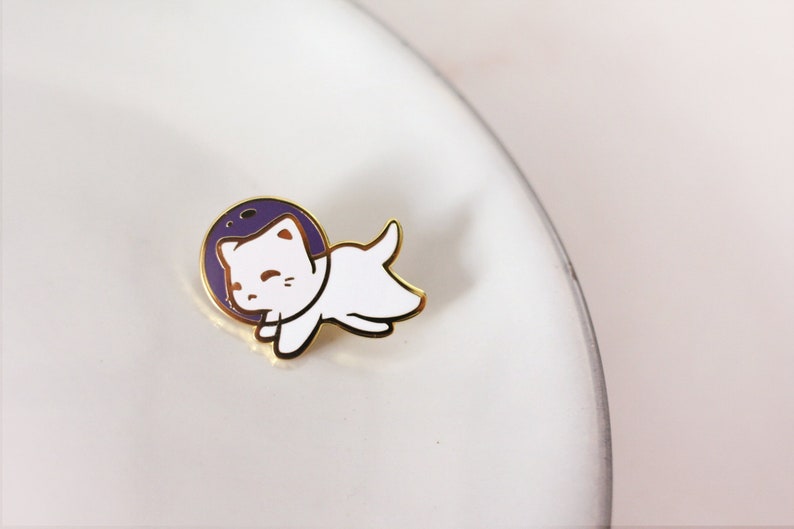 Puede incluir: Pin de esmalte dorado con un gato blanco que lleva un casco de astronauta morado. El gato est&aacute; en posici&oacute;n flotante con las patas extendidas.