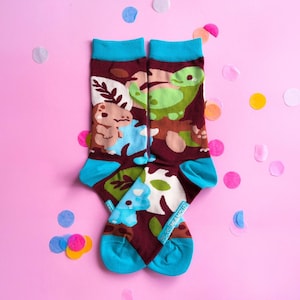 Könnte beinhalten: Ein Paar Socken mit dunkelweinroter Grundfarbe und einem bunten Dinosaurier-Muster, das hellgrüne, blaue und braune Comic-Dinosaurier sowie Blattmotive zeigt. Die Socken haben leuchtend türkisfarbene Bündchen, Fersen und Zehenbereiche.