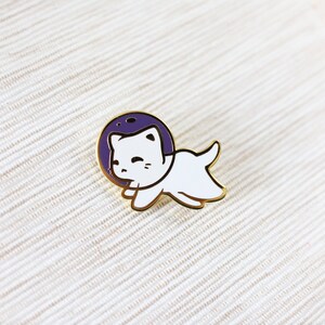Puede incluir: Un pin de esmalte dorado con un gato blanco que lleva un casco de astronauta morado. El gato est&aacute; en una pose de carrera con la boca abierta.