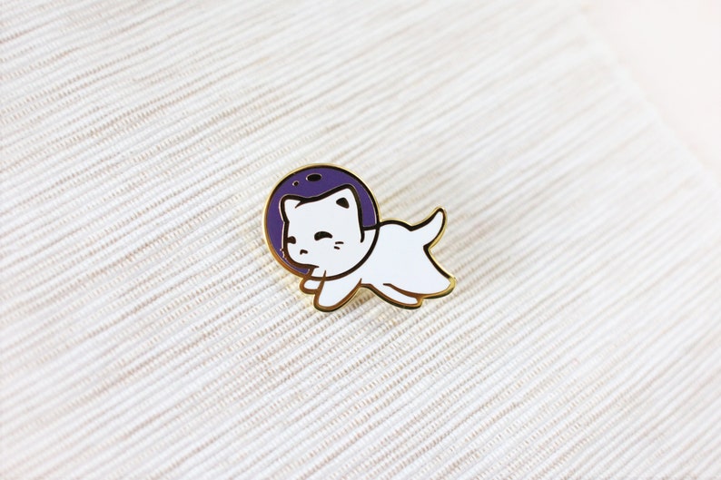 Puede incluir: Un pin de esmalte dorado con un gato blanco que lleva un casco de astronauta morado. El gato est&aacute; en una pose de carrera con la boca abierta.