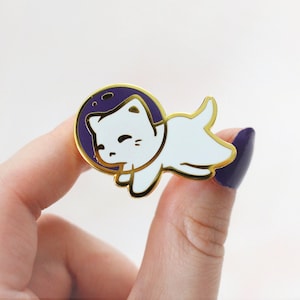 Puede incluir: Pin de esmalte dorado con un gato blanco que lleva un traje de astronauta morado, flotando en el espacio. El gato tiene una expresi&oacute;n feliz y mira hacia la derecha.