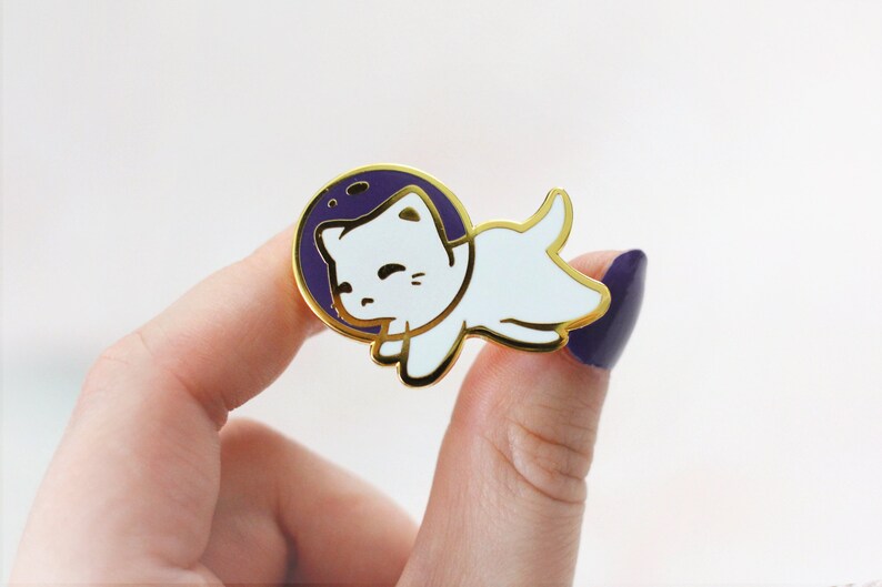 Puede incluir: Pin de esmalte dorado con un gato blanco que lleva un traje de astronauta morado, flotando en el espacio. El gato tiene una expresi&oacute;n feliz y mira hacia la derecha.