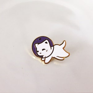 Puede incluir: Un pin de esmalte dorado con un gato blanco que lleva un casco de astronauta. El gato flota en un c&iacute;rculo morado, que se asemeja a un planeta o una luna.