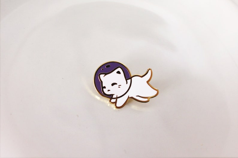 Puede incluir: Un pin de esmalte dorado con un gato blanco que lleva un casco de astronauta. El gato flota en un c&iacute;rculo morado, que se asemeja a un planeta o una luna.