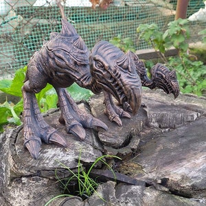 Puede incluir: Escultura detallada de criatura de color bronce. La escultura, con superficie texturizada, está posada sobre un tocón de árbol desgastado. La criatura tiene garras afiladas y una cabeza puntiaguda.