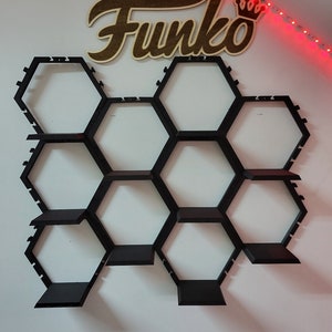 Puede incluir: Una estanter&iacute;a hexagonal negra con nueve estantes, montada en una pared blanca. Las estanter&iacute;as est&aacute;n dise&ntilde;adas para contener figuras Funko Pop. La palabra "Funko" est&aacute; escrita en letras de madera marr&oacute;n sobre las estanter&iacute;as.