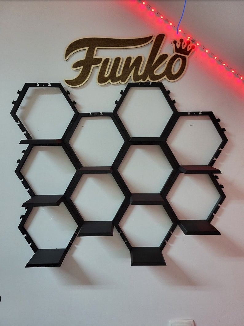 Puede incluir: Una estanter&iacute;a hexagonal negra con nueve estantes, montada en una pared blanca. Las estanter&iacute;as est&aacute;n dise&ntilde;adas para contener figuras Funko Pop. La palabra "Funko" est&aacute; escrita en letras de madera marr&oacute;n sobre las estanter&iacute;as.