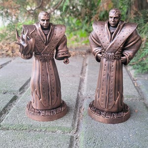 May include: Two bronze painted figurines of a person wearing a long robe with intricate details. The figures are standing on a base with a textured surface.