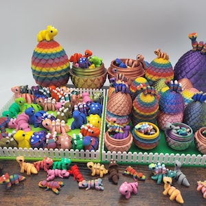 Może przedstawiać: Kolekcja kolorowych, jajowatych pojemników w kształcie smoka z małymi figurkami smoków w środku. Pojemniki są wykonane z materiału w kolorze tęczy i mają łuskowatą fakturę. Figurki są wykonane w różnych kolorach i ułożone w różnych pozach.