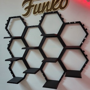 Puede incluir: Estantes hexagonales negros montados en una pared blanca, con un letrero de madera que dice "Funko" sobre los estantes.