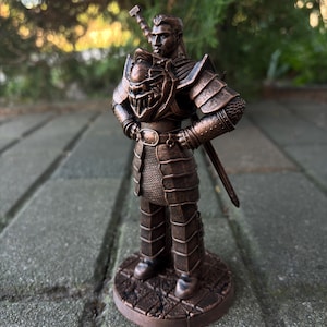 Op de afbeelding: Een bronskleurig beeldje van een krijger die op een ronde voet staat. Het figuur draagt een gesegmenteerd harnas, een riem met een gesp en een borststuk met een sculptuur van een monstergezicht. Een zwaard is op de rug bevestigd.