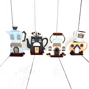 May include: Four whimsical coffee-themed decorations. The first is a light blue espresso maker, followed by a black teapot, a white kettle, and a tan mug. Each has a house-like design with windows and doors. Brown coffee beans are scattered around the black and white pieces.
