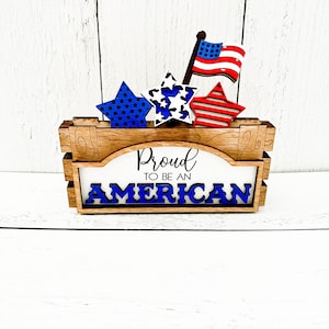 May include: Wooden sign with the text "Proud to be an American" in blue letters. The sign is decorated with three red, white, and blue stars and a small American flag.