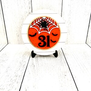 May include: A white round wooden sign with a black spider and web design on an orange background. The number 31 is in the center of the sign with black bats on either side. The sign is on a white wooden stand.