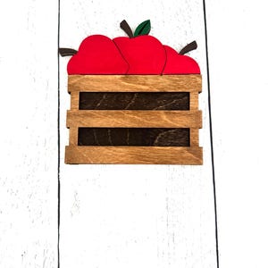Puede incluir: Caja de madera llena de tres manzanas rojas con hojas verdes y tallos marrones. La caja es de madera marr&oacute;n claro con listones marrones oscuros. El fondo es una superficie de madera blanca.