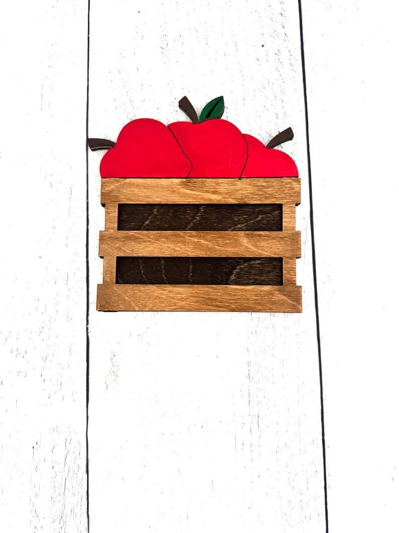 Puede incluir: Caja de madera llena de tres manzanas rojas con hojas verdes y tallos marrones. La caja es de madera marr&oacute;n claro con listones marrones oscuros. El fondo es una superficie de madera blanca.