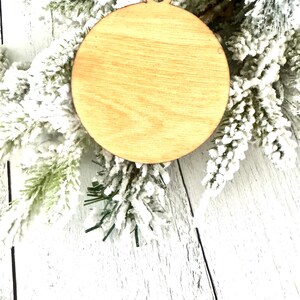 Puede incluir: Un adorno de madera redondo y liso con una cuerda roja atada a la parte superior. El adorno est&aacute; sobre una superficie blanca y nevada.