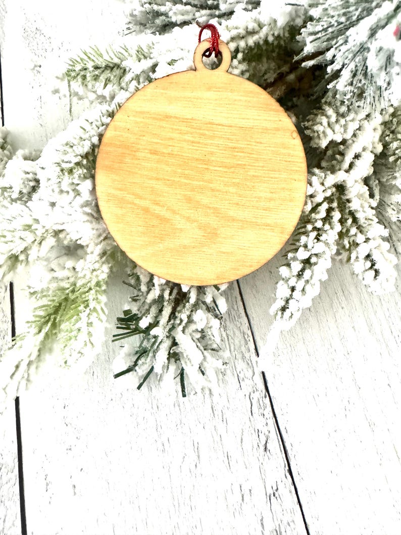 Puede incluir: Un adorno de madera redondo y liso con una cuerda roja atada a la parte superior. El adorno est&aacute; sobre una superficie blanca y nevada.